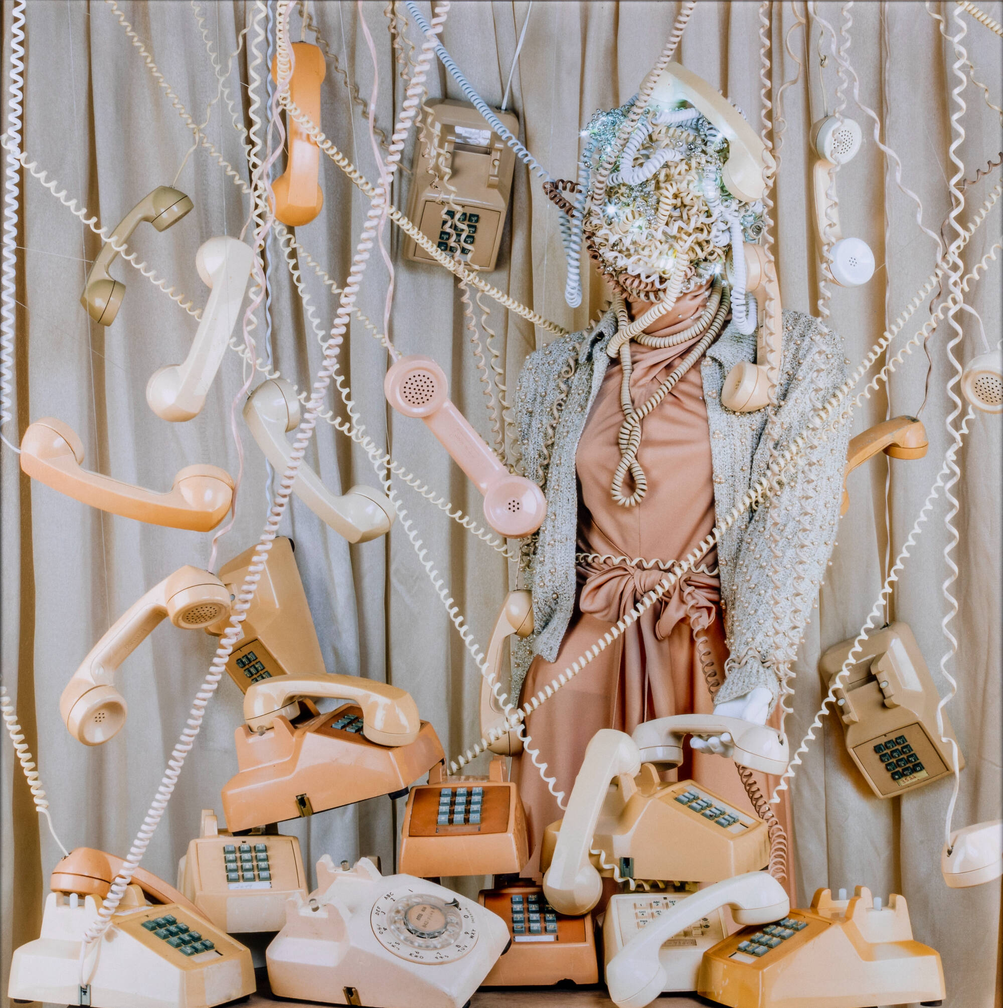 Color photograph of a female figure, face covered, surrounded by telephones and cords.